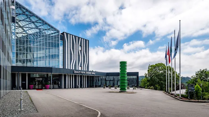 A modern hotel building displaying "QUALITY HOTEL EDVARD GRIEG" has glass facades with abstract patterns. Flagpoles and a green sculpture stand outside against a partly cloudy sky.