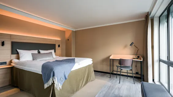 A neatly made bed with a blue throw blanket lies beside a wooden desk and chair, illuminated by a lamp. Soft, earthy tones decorate the room with natural light streaming in.