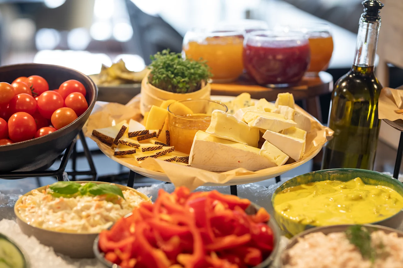 Ost- och grönsaksfat ordnat med skivade ostar, cocktailtomater, röd paprika och olika dippsåser. En olivoljeflaska och små burkar med konserver syns i en ljus matsalsmiljö.