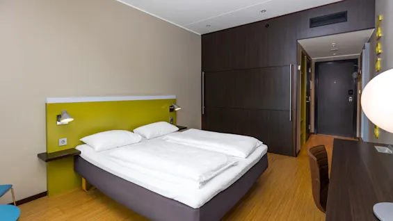 A neatly made double bed with white linens stands in a modern hotel room, featuring wooden flooring, a green headboard, a desk with a lamp, and a closed dark door.