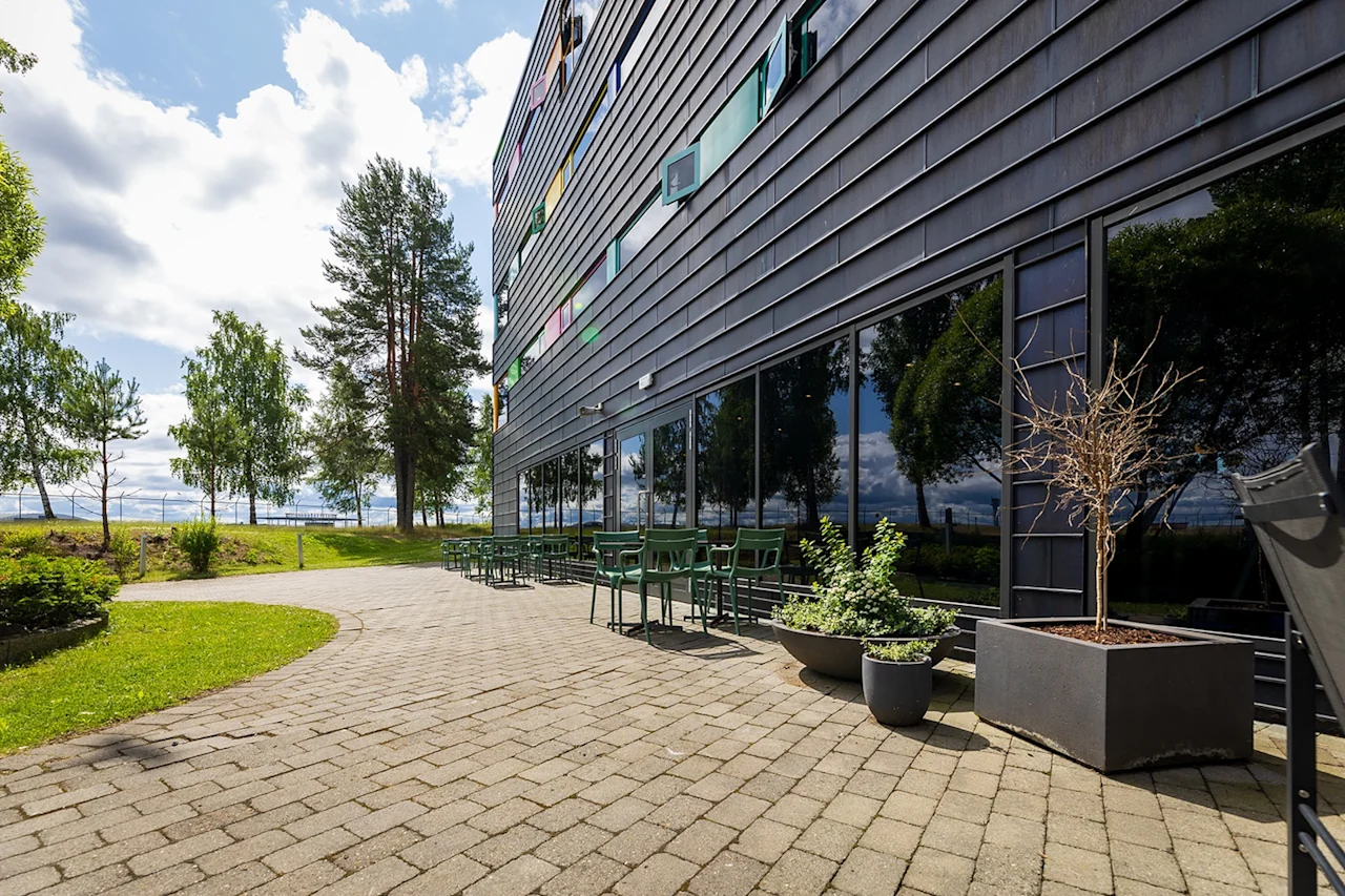 Byggnad med reflekterande glasfönster och gröna stolar på en stenlagd uteplats som sträcker sig mot ett gräsområde med träd under en molnig himmel.