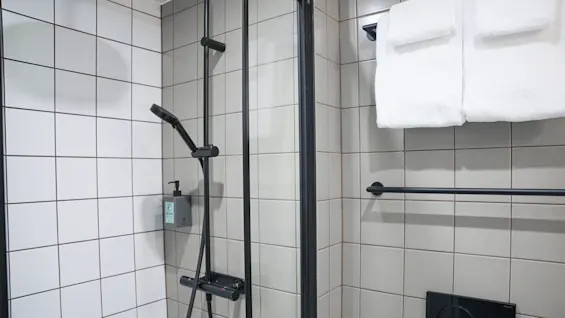 Dusjhode montert på flislagt baderomsvegg, med en såpedispenser merket "ALL-IN-ONE." Nær ved er et svart håndklekroker med to foldede hvite håndklær.