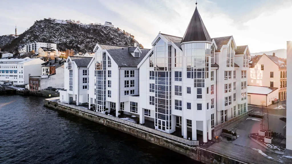 A large white building with pointed roofs reflects on calm water, set against snowy hills and adjacent urban structures under a blue sky.