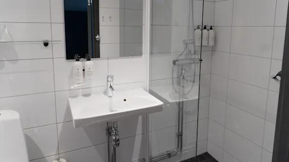 En vit handfat med kran är monterad på en kaklad badrumsvägg. Ovanför reflekterar en spegel en del av duschområdet med monterade flaskor. Toalettpapper står i närheten.