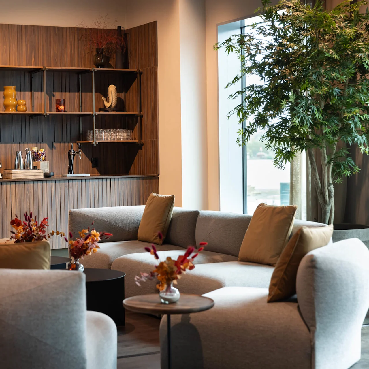 Kaffemaskin står på en trädisk i en modern lounge, omgiven av mjuka grå sittplatser och dekorativa växter, vilket skapar en mysig atmosfär.