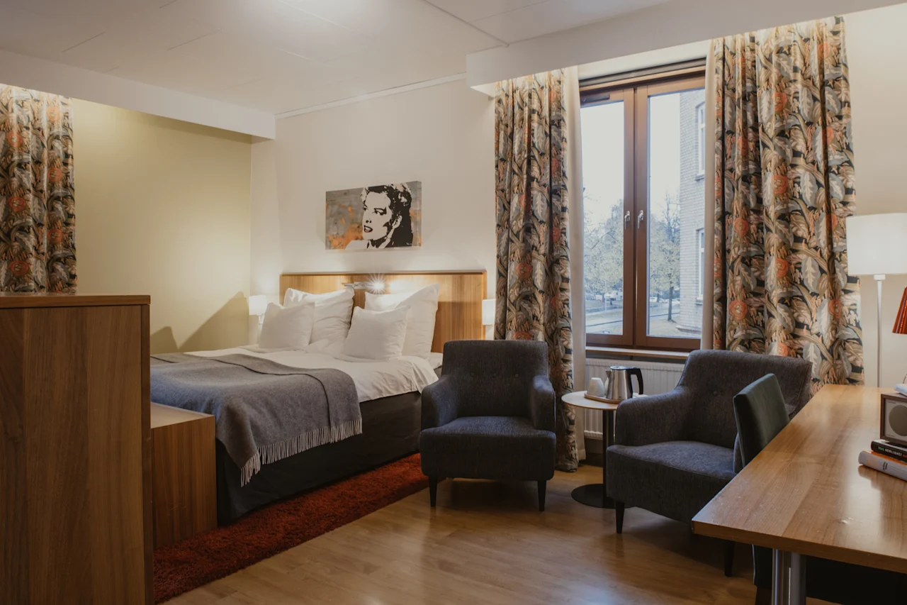 A cozy hotel room at Home Hotel Kung Oscar with a comfortable bed, two armchairs, a desk, and a window with patterned curtains.