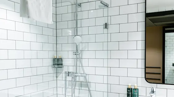 Bruser med hvide fliser, holder et håndklæde og brusehoved, ved siden af en vask med toiletsager, alt i et lyst, moderne badeværelse.