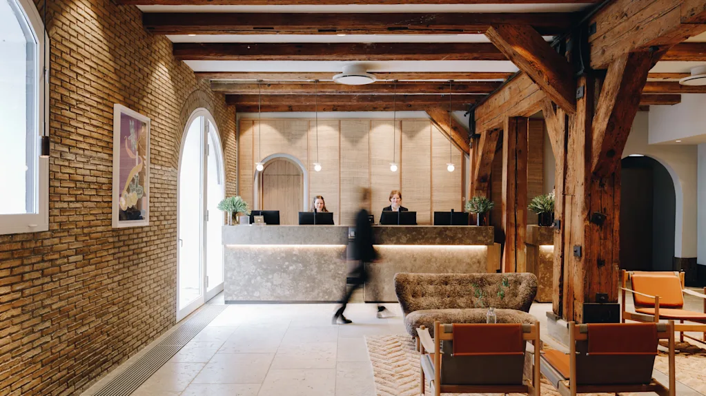 En person går gjennom en hotellobby med resepsjon, ansatte, murvegger og trebjelker, noe som skaper en varm atmosfære.