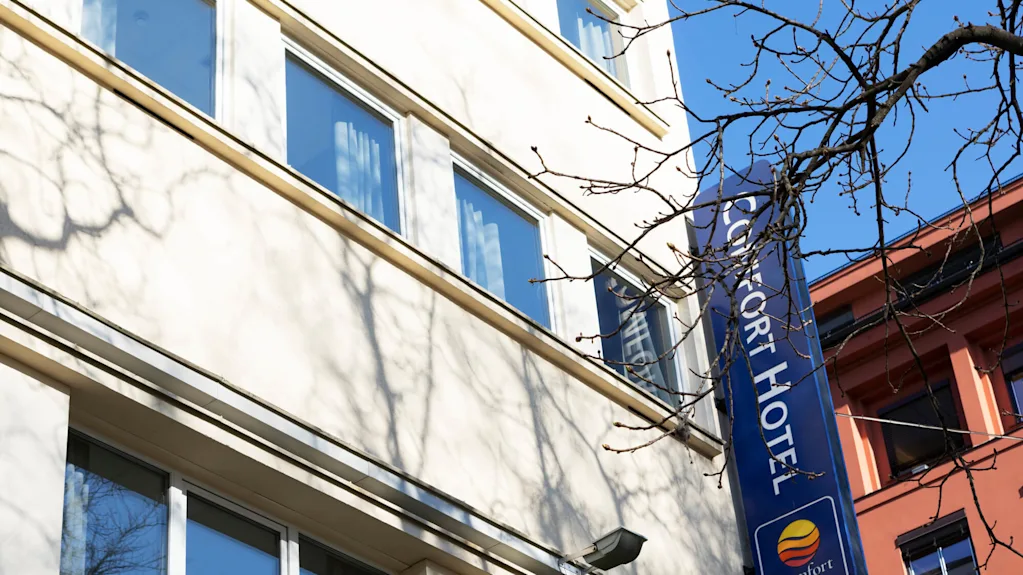 Hotellikyltti on kiinnitetty rakennukseen nimellä ja logolla, lehdettömän puun ja kirkkaansinisen taivaan välissä.