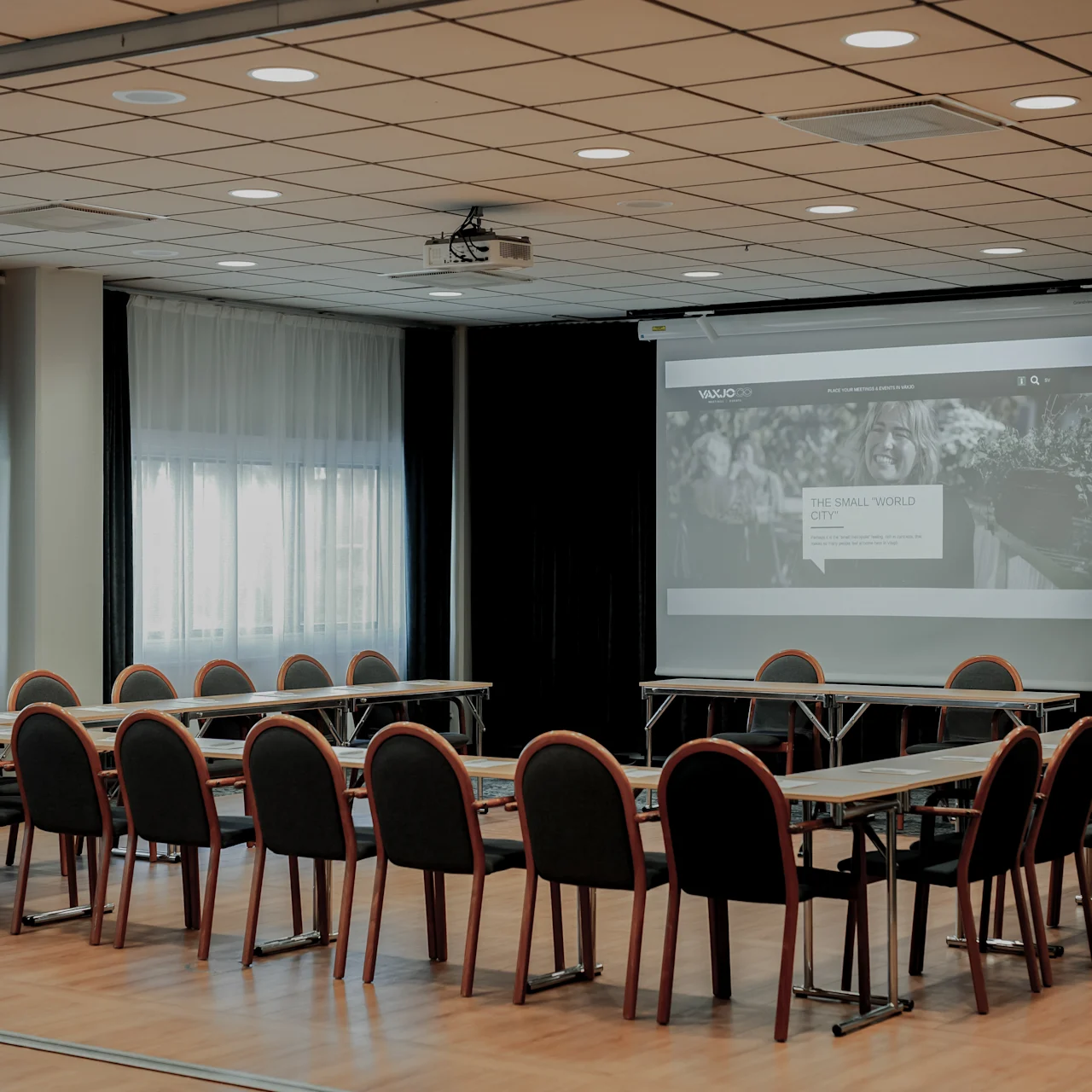 Two rows of conference chairs face a projector screen displaying content in a well-lit meeting room with wood flooring and large windows.