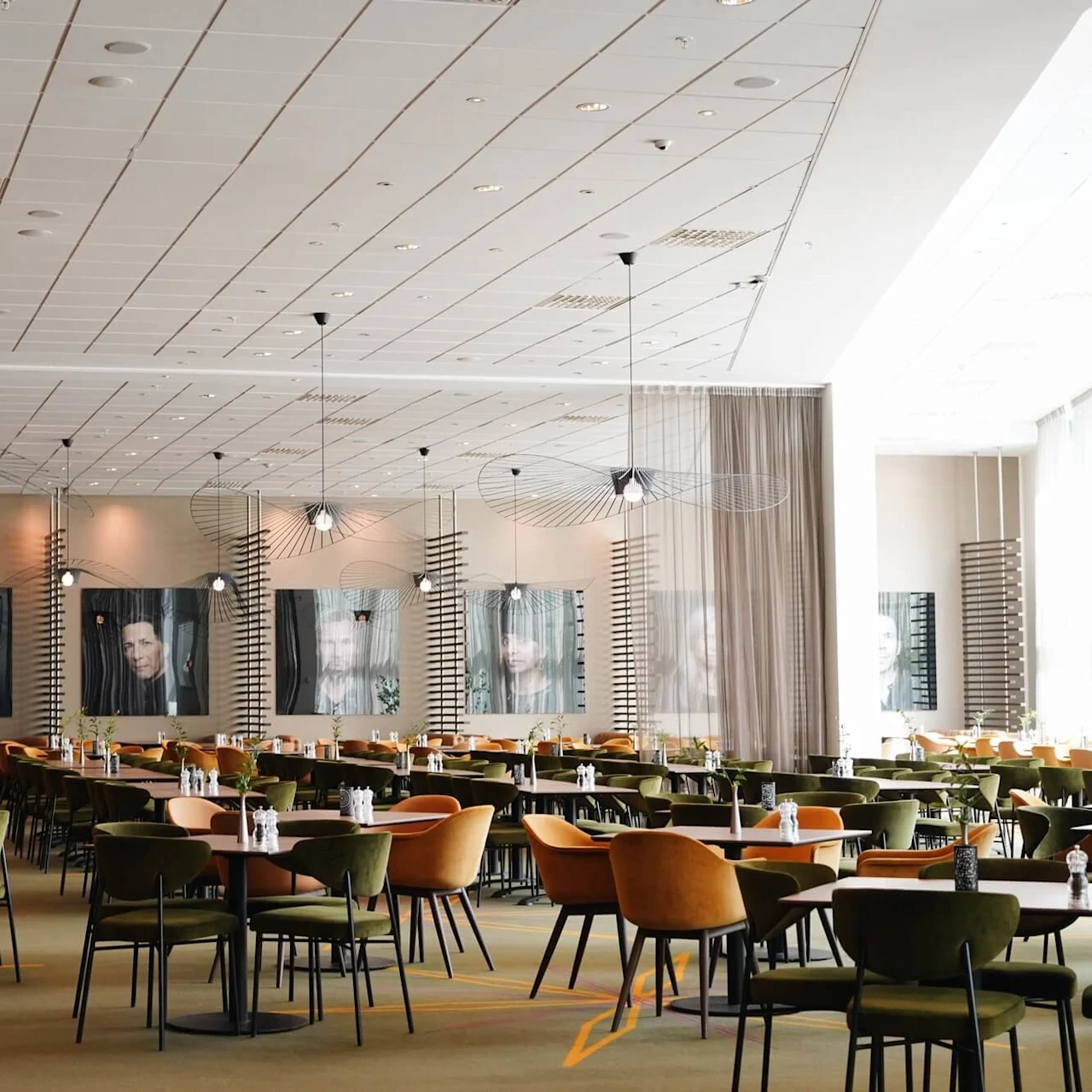 A spacious dining area features rows of tables and colorful chairs. Decorative curtains hang from the ceiling. Walls are adorned with large, framed portraits. Natural light fills the room.