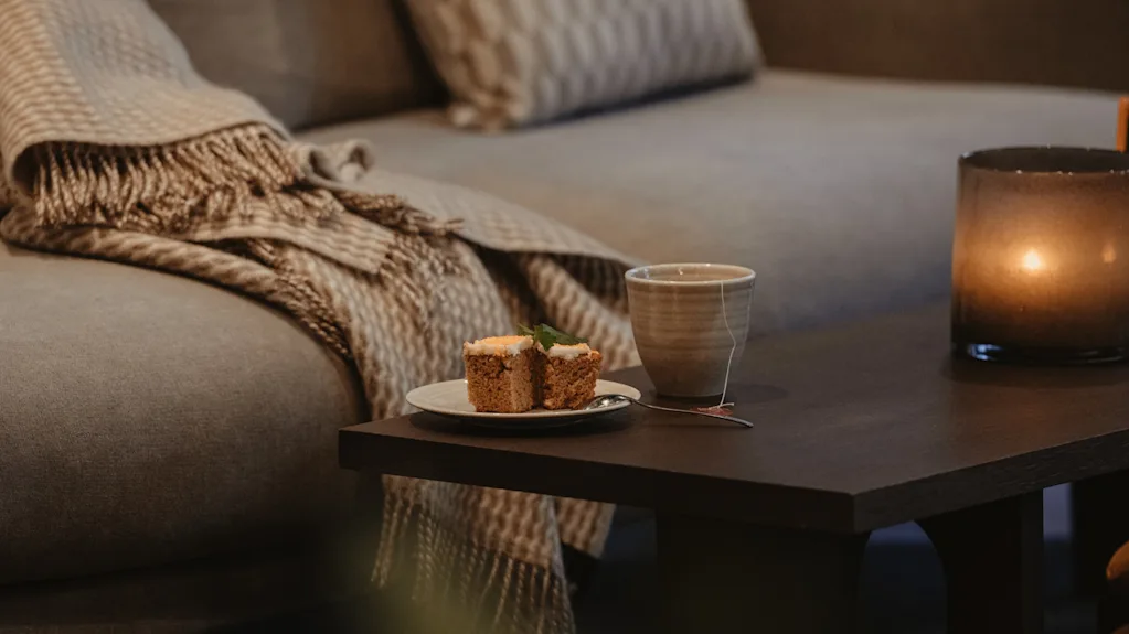 En koselig scene med et teppe på en sofa, et sidebord med kake, te og et varmt, glødende lys.