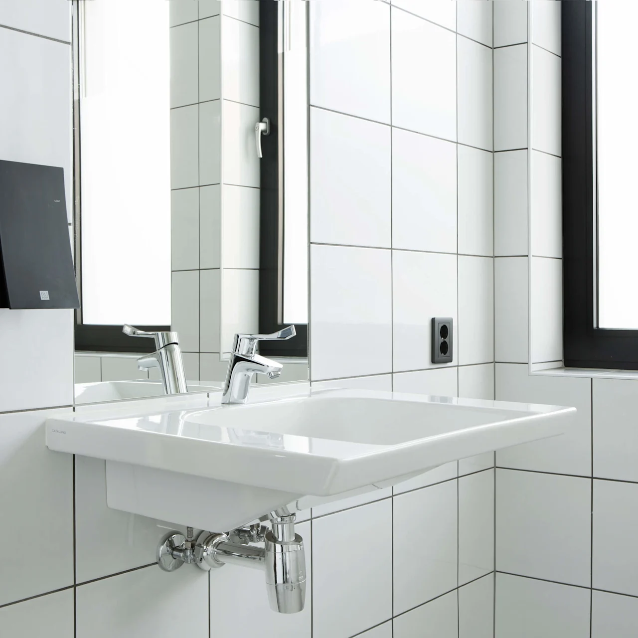 A white ceramic sink with dual chrome faucets is mounted on a tiled bathroom wall near a large frosted window. A black booklet hangs on the wall beside the sink.