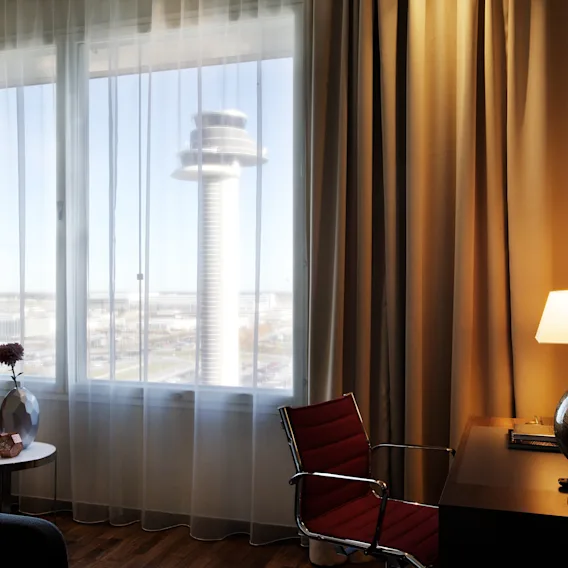 Et lyst hotellrom med et stort vindu som gir en fremtredende utsikt over et flyplasstårn, sammen med et skrivebord og en komfortabel stol.