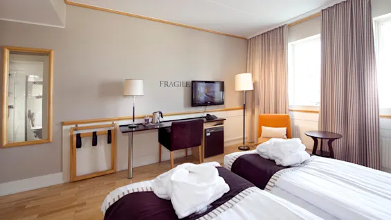 Ett snyggt hotellrum med två sängar, ett skrivbord med en TV och en spegel som reflekterar ett badrum.