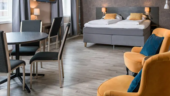 A modern hotel room features a bed with yellow cushions, a small round dining table with chairs, and two orange armchairs. Gray curtains and a mounted TV complete the setting.
