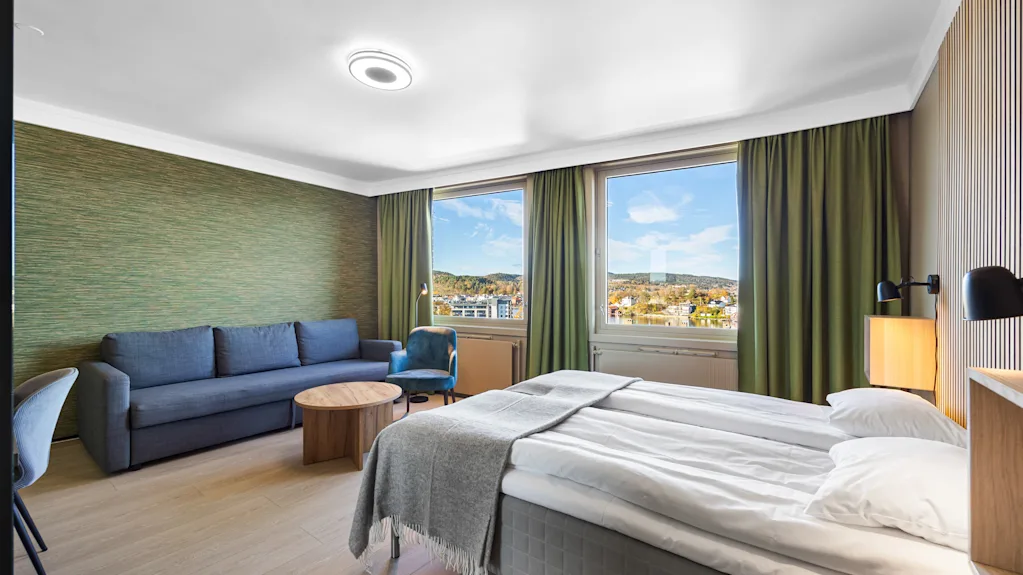 Lyst hotellrom med dobbeltseng, sofa og vinduer med utsikt.