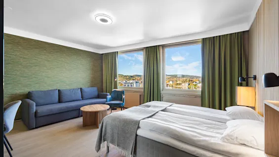 Lyst hotellrom med dobbeltseng, sofa og vinduer med utsikt.