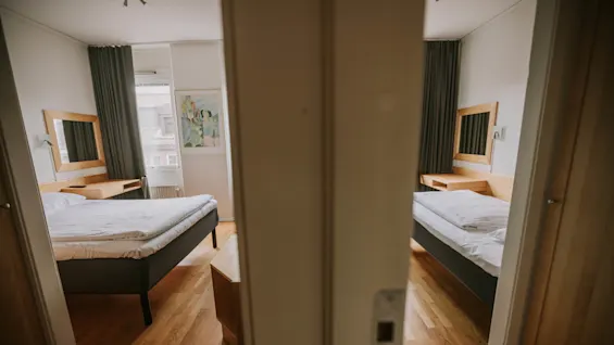 To tilstødende soveværelser med trægulve, hver med en pænt redt seng og et skrivebord med spejl. Mørke gardiner pryder vinduerne, og kunstværker hænger på den fælles væg.