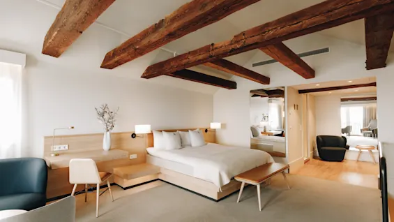 Ljust hotellrum med synliga takbjälkar i trä, en stor säng och modern minimalistisk inredning.