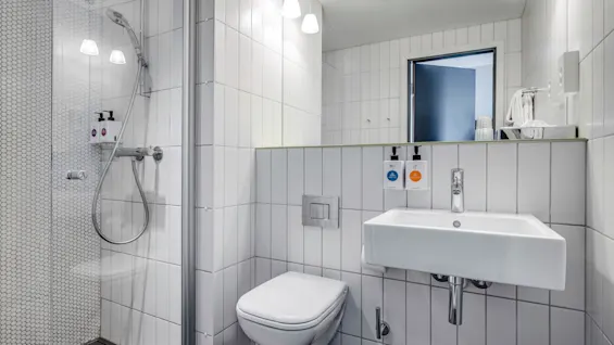 Ett badrum har ett väggmonterat handfat med spegel ovanför, en toalett och en dusch med glasväggar. Väggarna har vita kakelplattor. Tvålpumpar monterade nära handfatet.