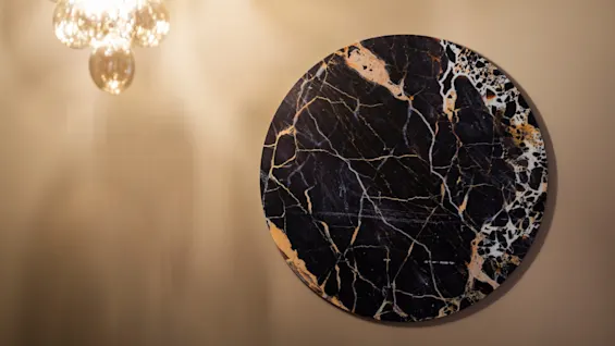 A circular black marble art piece with gold and white veins hangs on a beige wall at Home Hotel Grand Bodø, with a light fixture above.