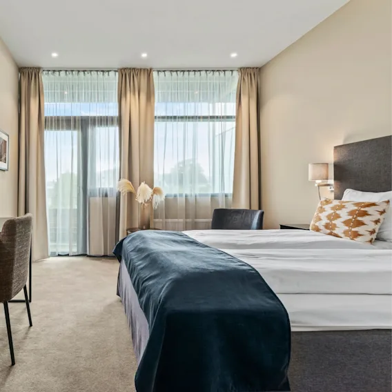 Lyst og moderne hotellrom med en oppredd dobbeltseng, store vinduer og et behagelig sitteområde.