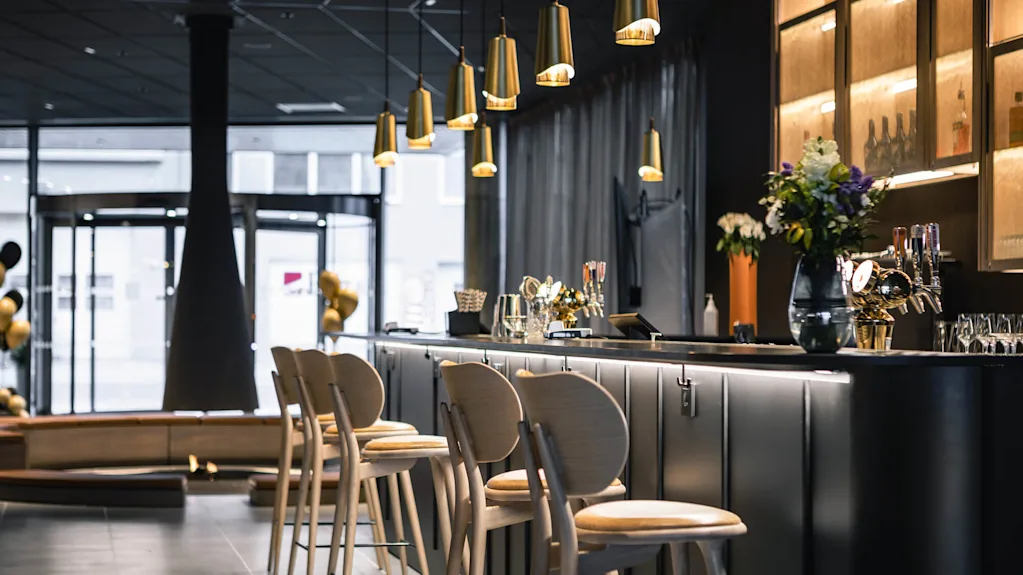 Fyra barstolar i trä står vid en elegant, belyst bar i en modern restaurang. Ovanför hänger gyllene hänglampor, vilket ger värme till den stiliga interiören.