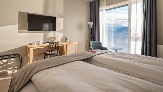 Et komfortabelt hotelværelse med en stor seng, et skrivebord med TV og en fantastisk udsigt over en sø og bjerge fra vinduet.