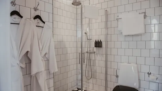 Två vita badrockar hänger på krokar bredvid en glasdusch. I duschen står svarta flaskor på en hylla. En toalett står i närheten, omgiven av vita kakelväggar.