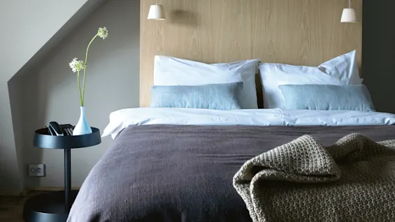 A bed with white pillows and blue cushions stands against a wooden headboard. A side table holds a vase with flowers and a phone, beneath hanging lamps.