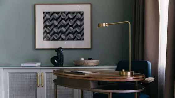 Et trebord med en gullampe står foran et skap, under et innrammet geometrisk kunstverk. Myke gardiner henger ved siden av bordet, og skaper en koselig atmosfære.