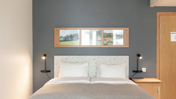 A comfortable hotel room featuring a bed with white linens, a speckled headboard, and wall-mounted lamps. Landscape art adorns the wall.