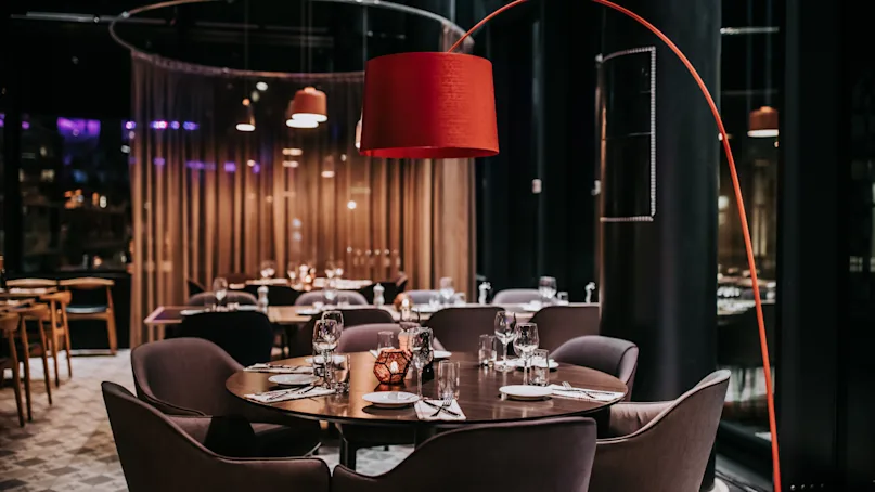 A round dining table set for a meal, surrounded by cushioned chairs. Overhead, a large red lamp provides light. The setting is a dimly lit, modern restaurant.