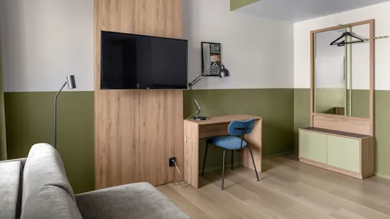 En väggmonterad TV vetter mot ett litet skrivbord av trä med en blå stol och svart lampa, belägen i ett rum med gröna och gräddfärgade väggar och en stor spegel med klädhängare.