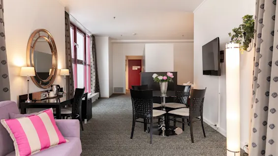 Hyvin varusteltu hotellihuone, jossa on erillinen oleskelutila, ruokapöytä ja suuret ikkunat, luoden kutsuvan tunnelman.