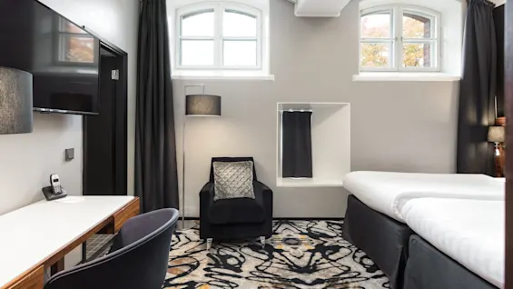 A modern hotel room with twin beds, a black armchair, a desk, curtains, and a patterned carpet. The decor is in neutral tones, with a large window letting in natural light.