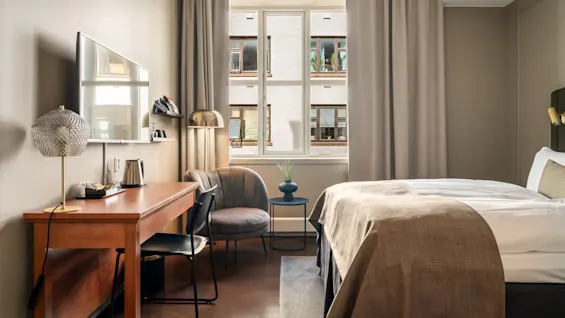 Et pænt arrangeret hotelværelse med seng og skrivebord ved et stort vindue, i neutrale toner. Indstillingen er hyggelig og moderne, hvilket antyder et komfortabelt ophold. Ingen tekst er til stede.