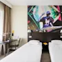 Two single beds in a hotel room, topped by a mural of a DJ performing, with a desk and a window adorned with brown curtains.