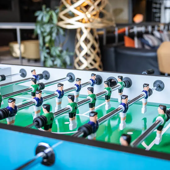 Foosball table with figures positioned for play, in a lounge area featuring modern furniture and decorative lighting structures, creating a relaxed, recreational atmosphere.