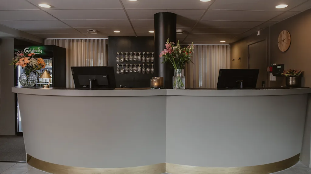 Reception med två bildskärmar och en vas med blommor. En Carlsberg-dryckkylen står i närheten. Vinglas syns på ett ställ mot väggen.