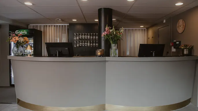 Resepsjonsskranke med to skjermer og en vase med blomster. En Carlsberg-drikkeavkjøler står i nærheten. Vinglass er synlige på en hylle mot veggen.