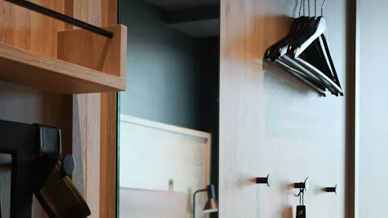 Wooden panel with hanging black coat hangers and a shoehorn is mounted beside a mirror. A neatly made bed with white pillows is visible in the adjacent room.