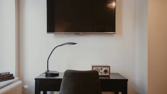 Hotelværelses skrivebord med TV ovenover. Lampe, radio og bøger med titlen FALSKY SPEL er på eller nær skrivebordet.