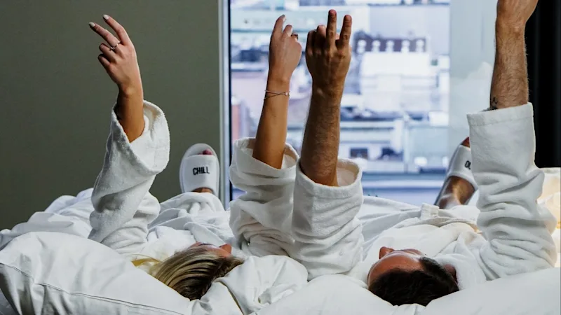 To personer i hvide badekåber ligger i sengen med armene hævet, overfor et vindue der viser et bylandskab.