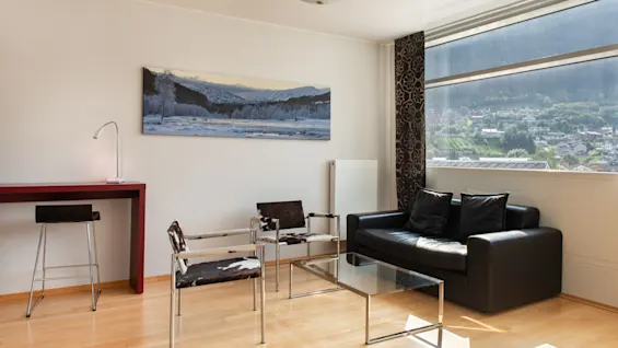 En moderne stue med en sort sofa, stole med kohud og et stort vindue med udsigt over en by og bjerge.
