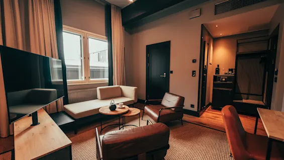 A modern hotel room with a comfortable seating area, a large window, and warm lighting, creating a welcoming atmosphere.