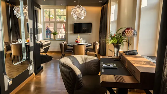 Et stilfullt hotellrom har et skrivebord i tre med en mønstret stol, dekorativ lampe og planter, vendt mot et spiseområde med moderne stoler og en lysekrone.