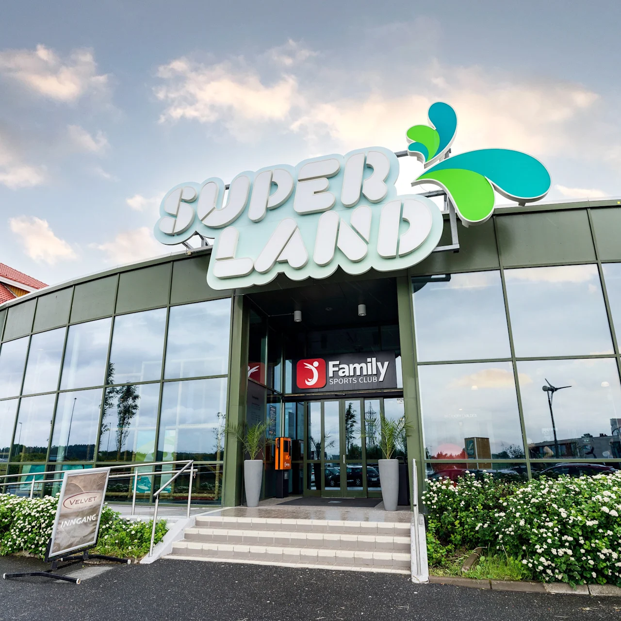A curved glass building entrance with "SUPER LAND" sign above. Below, a "Family Sports Club" sign by stairs, surrounded by greenery. Sign reads: "VELVET INNGANG."