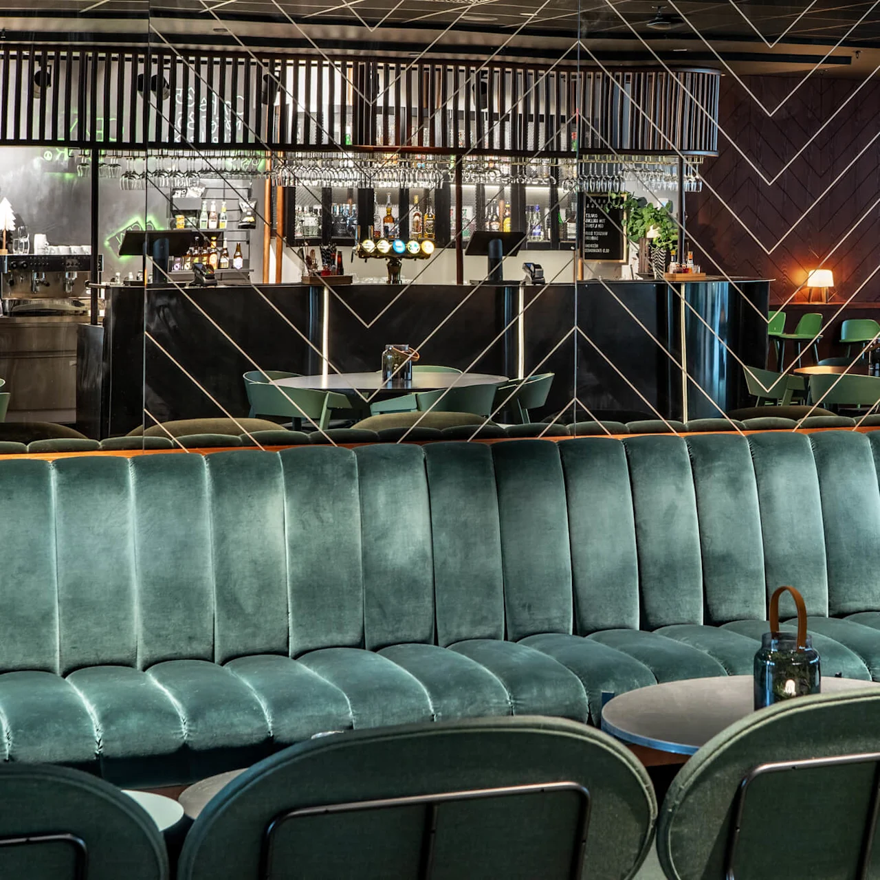 A large, elegant green velvet sofa faces a stylish bar area, featuring hanging glassware and ambient lighting, surrounded by green chairs and tables in a modern lounge setting. 